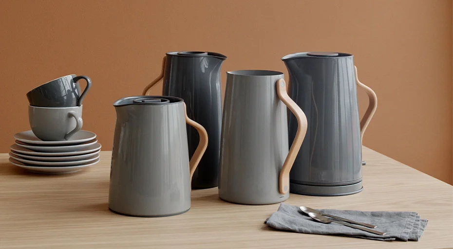 Mehrere graue Stelton Wasserkocher, Kannen und Tassen auf einem Holztisch vor einer braunen Wand, mit Besteck und Servietten.