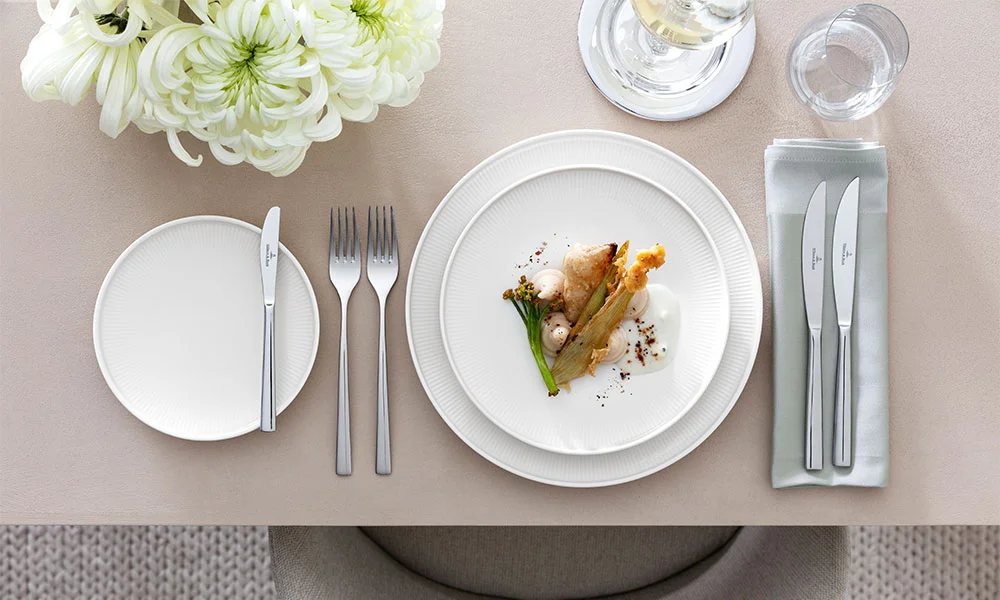 Eleganter Tisch mit weißem Porzellangeschirr der Serie Afina von Villeroy &amp; Boch, Besteck, Blumendekoration und angerichtetem Gourmetgericht.