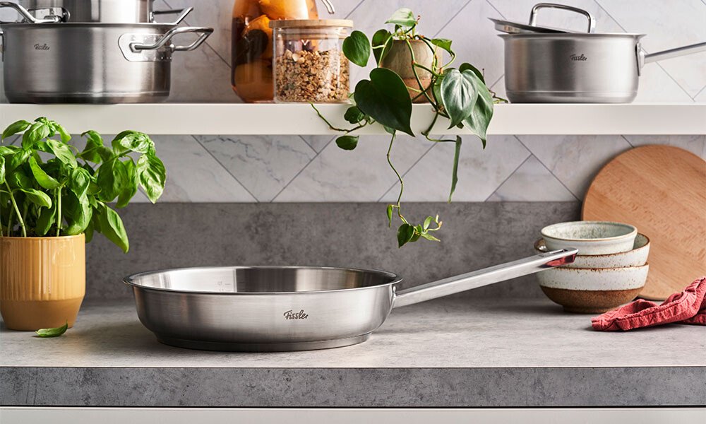 Pfanne von Fissler aus Edelstahl auf Küchenarbeitsplatte, umgeben von Pflanzen, Schüsseln und Töpfen vor einer gefliesten Rückwand.