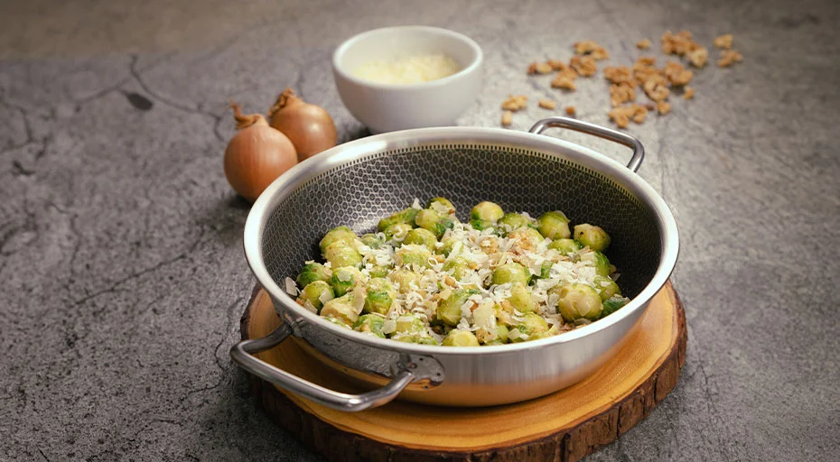 Servier-Pfanne der Serie Vulcano Cut Resist Pure von Spring mit Rosenkohl und geriebenem Käse, dekoriert mit Zwiebeln und Haferflocken, auf Holzbrett serviert.