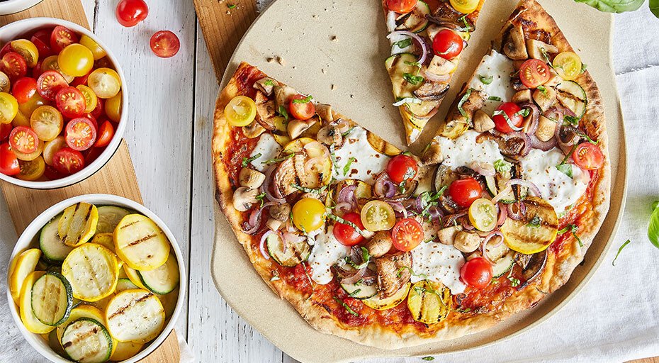 Pizza mit Gemüsetoppings auf Steinplatte, umgeben von Schalen mit Kirschtomaten und gegrilltem Gemüse.