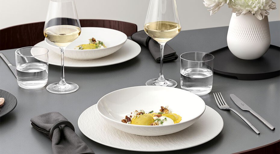Eleganter gedeckter Tisch mit Tellern der Serie Manufacture Ice von Villeroy &amp; Boch mit Pasta, Weingläsern, Wassergläsern, dunklen Servietten und Blumen in Vase.