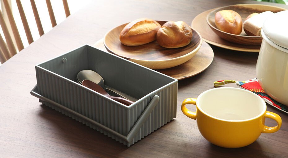 Eine Aufbewahrungsbox von Hachiman, befüllt mit Besteck, daneben Brötchen auf Holzplatten und eine gelbe Tasse auf einem Holztisch.