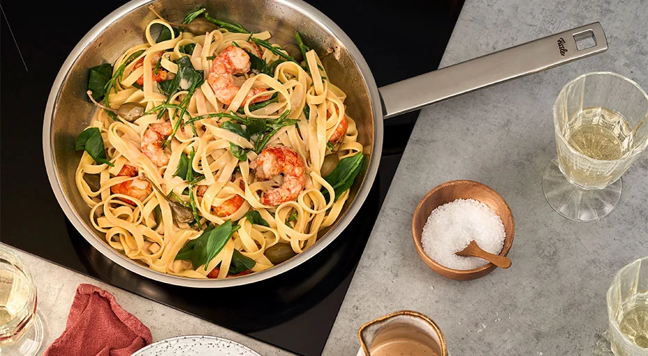 Fissler Pfanne mit Linguine, Garnelen, Rucola und Kirschtomaten; daneben Salzschale und Weinglas auf Arbeitsplatte.