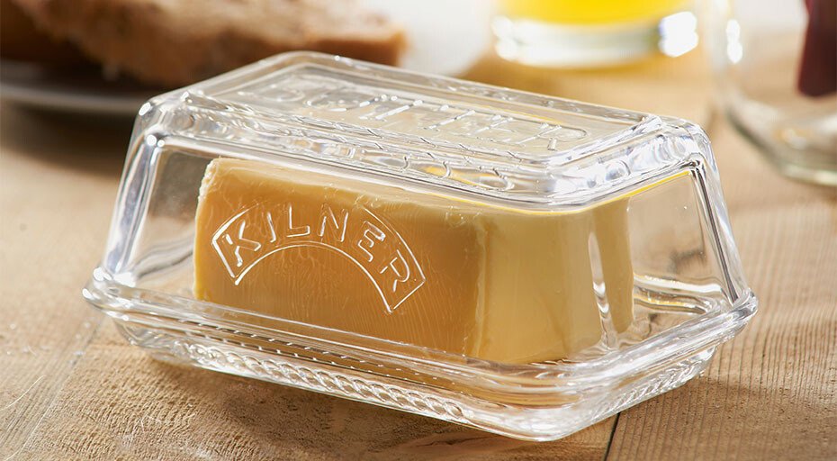 Butter in gläserner Butterdose von Kilner auf Holztisch. Hintergrund unscharf mit Brot und Getränk.