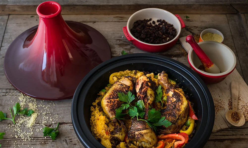 Traditionelle marokkanische Tajine von Le Creuset mit Hähnchen, Paprika und Gewürzen, garniert mit Petersilie, serviert auf einem Holztisch.