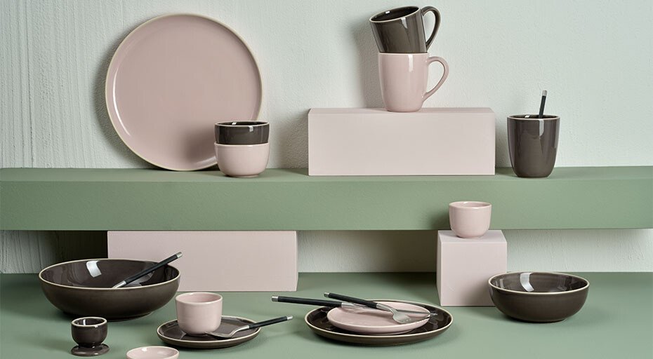 Keramikgeschirr in Rosa und Grau auf gestaffelten grünen Regalen, bestehend aus Tellern, Tassen und Schalen.