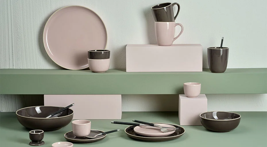 Keramikgeschirr in Rosa und Grau auf gestaffelten grünen Regalen, bestehend aus Tellern, Tassen und Schalen.