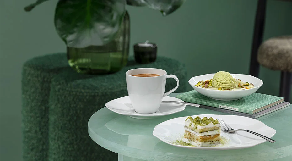 Villeroy &amp; Boch Pura Tasse mit Kaffee, Teller und Schale mit Dessert auf Glastisch vor grünem Hintergrund.