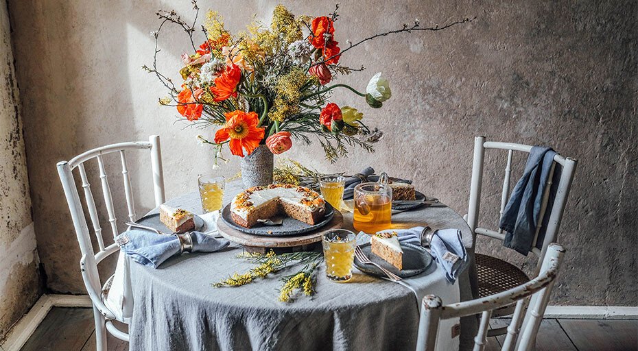 Ein rustikal gedeckter Tisch mit blau-grauer Tischdecke, Blumenstrauß, Kuchen und Getränken, umgeben von weißen Stühlen.