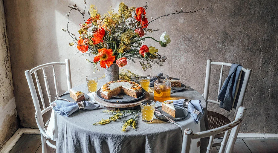 Ein rustikal gedeckter Tisch mit blau-grauer Tischdecke, Blumenstrauß, Kuchen und Getränken, umgeben von weißen Stühlen.