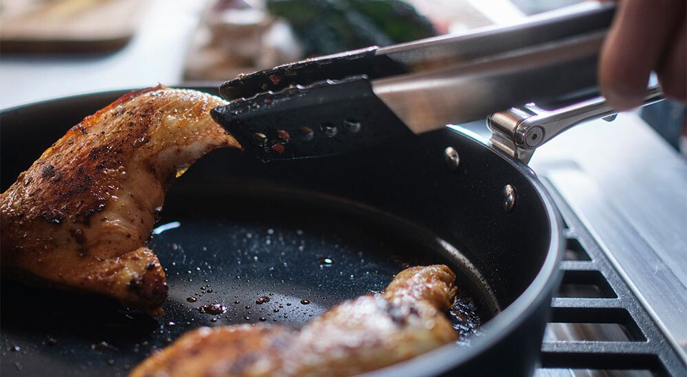 Nahaufnahme von Hähnchen in einer Keramik-Pfanne, das mit einer Metallzange gewendet wird.