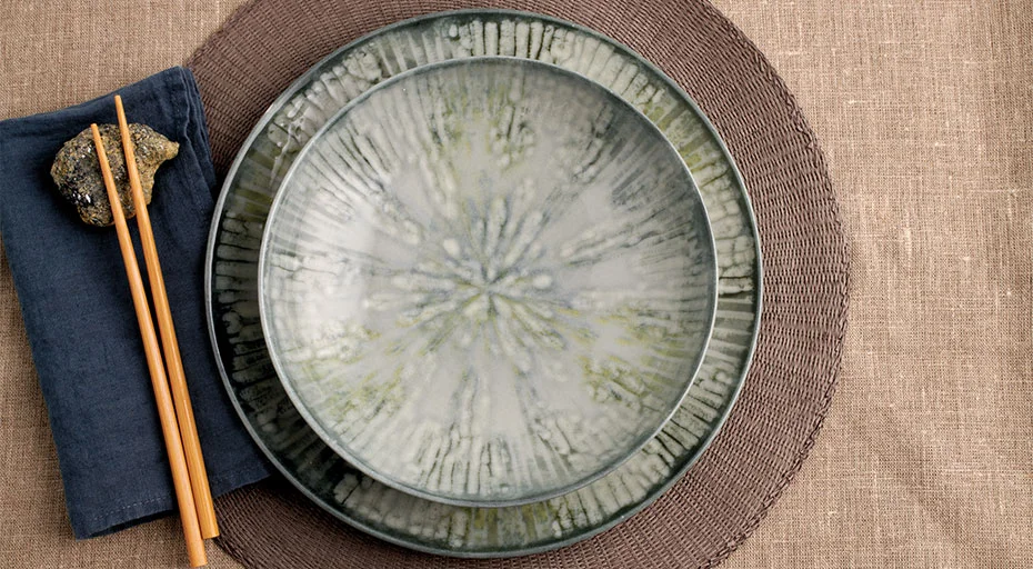 Rosenthal Arthur Krupp Keramikteller auf braunem Platzdeckchen, daneben Holzstäbchen auf dunkelblauer Serviette.