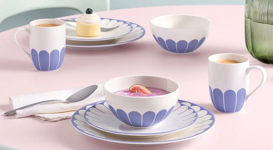 Tassen, Schalen und Teller der Serie Fleur Bleu von Villeroy &amp; Boch auf Frühstückstisch, serviert mit Joghurt, Tee und Gebäck auf rosa Tisch.