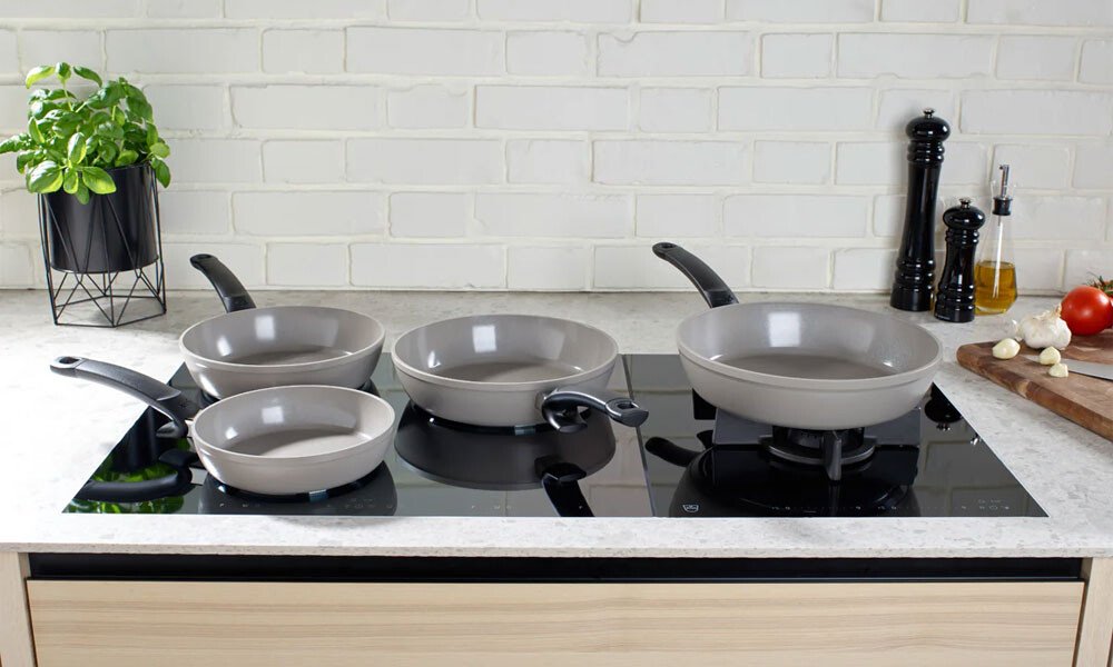 Fünf graue Pfannen der Serie Ceratal von Fissler auf einem modernen Induktionskochfeld in einer Küche, umgeben von Gewürzen und Kräutern.