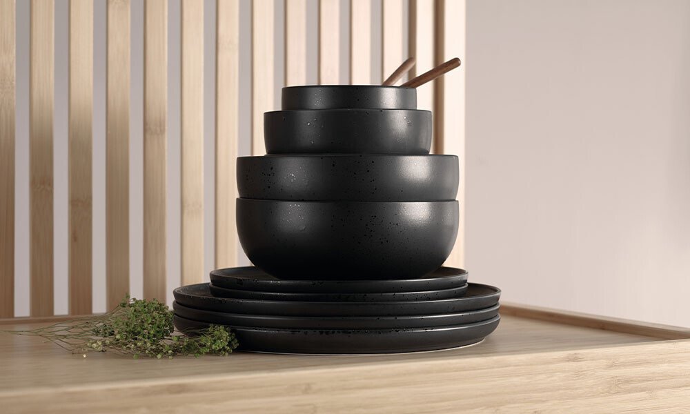 Schwarze, ineinander gestapelte Keramikschalen mit Löffeln auf Holzuntergrund, dekoriert mit Kräutern.