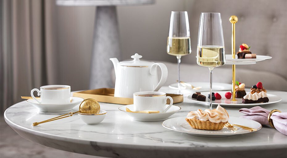 Edles Teeservice der Serie Château Septfontaines von Villeroy & Boch mit goldenen Löffeln auf Marmortisch, Sektgläser und Gebäck auf Etagere, elegante Atmosphäre.