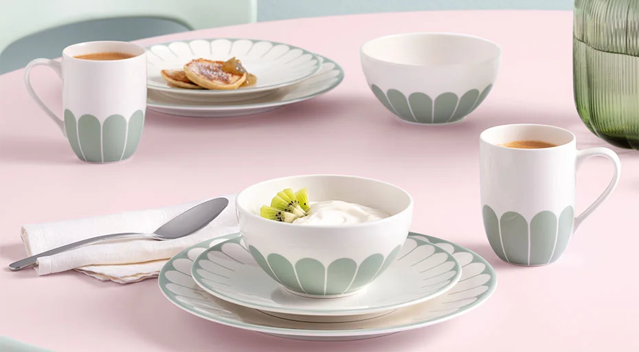 Geschirr-Set Villeroy &amp; Boch Fleur Vert auf Tisch: Teller, Tassen, Schüssel mit Joghurt und Kiwi, neben Kaffee und Pfannkuchen auf rosafarbenem Untergrund.