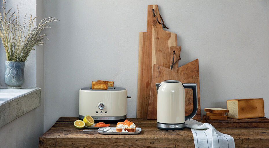 Toaster und Wasserkocher von KitchenAid auf Holztisch, daneben Schneidebretter, Kräutervase und Teller mit Speisen.