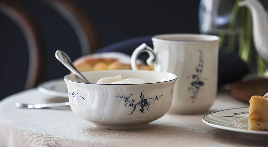 Porzellantasse und -schale der Serie Alt Luxemburg von Villeroy &amp; Boch mit blauem Blumenmuster auf gedecktem Tisch, Löffel in der Schale, im Hintergrund unscharf Gebäck.