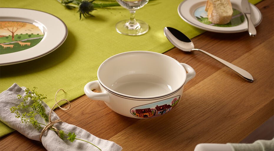 Tischgedeck mit Suppen-Tasse von Villeroy &amp; Boch, Teller, Besteck und grüner Tischdecke auf Holztisch. Deko mit Kräutern und gebundenem Stoffserviette.
