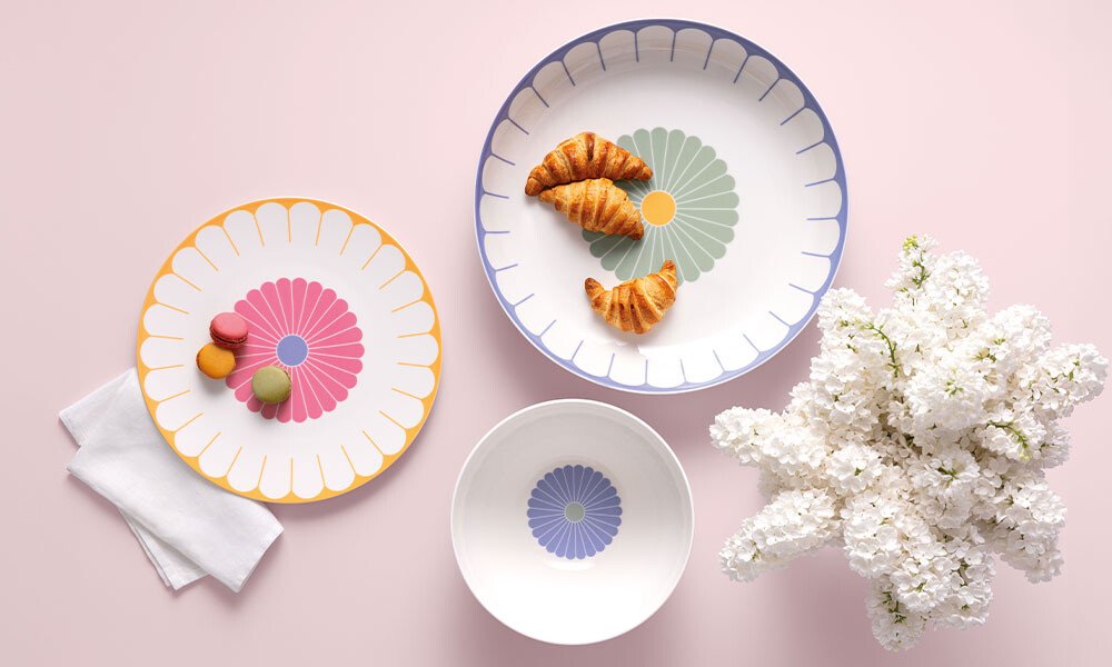 Porzellangeschirr der Serie Fleur von Villeroy &amp; Boch mit floralem Muster, serviert mit Croissants und Macarons. Daneben weiße Blumen auf rosa Hintergrund.