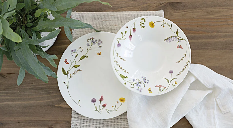 Zwei Porzellanteller von Hutschenreuther Nora Spring Vibes mit floralen Mustern auf Holztisch neben einer grünen Pflanze und einem weißen Tuch.