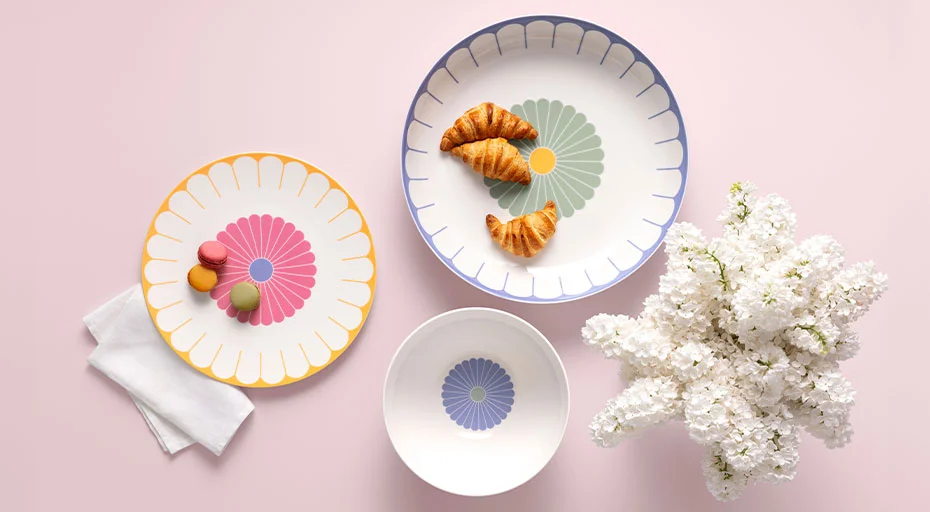 Teller der Serie Fleur Couleur von Villeroy &amp; Boch mit farbigen, floralen Mustern, Croissants und Macarons auf hellem Hintergrund, daneben weiße Blüten.