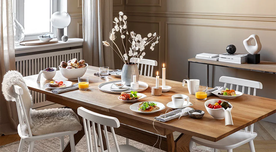 Geschirr der Serie Flow von Villeroy &amp; Tisch auf Holztisch mit sechs weißen Stühlen, gedeckt für Frühstück mit Saft, Brötchen, Lichtelementen und dekorativer Vase.