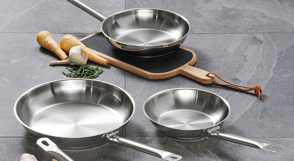 Edelstahlpfannen verschiedener Größen auf einem grauen Untergrund, umgeben von Gemüse.