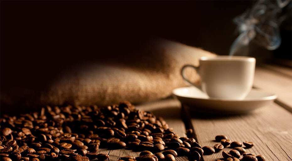 Kaffeetasse mit Dampf, umgeben von gerösteten Kaffeebohnen auf Holztisch. Hintergrund dunkel gehalten.