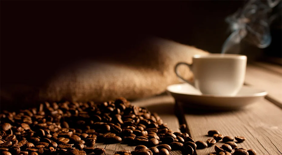 Kaffeetasse mit Dampf, umgeben von gerösteten Kaffeebohnen auf Holztisch. Hintergrund dunkel gehalten.