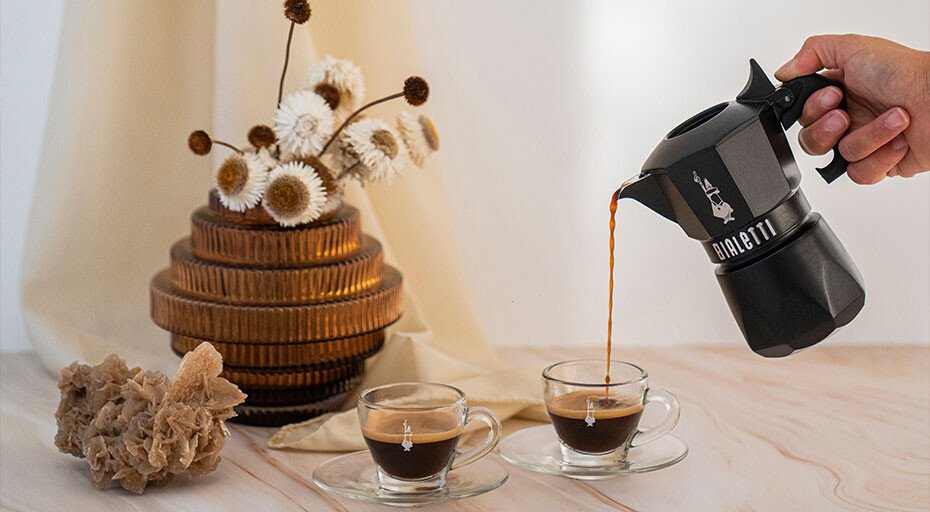 Eine schwarze Espressokanne von Bialetti gießt Espresso in zwei Gläser, daneben Trockenblumenarrangement auf hellem Holz.