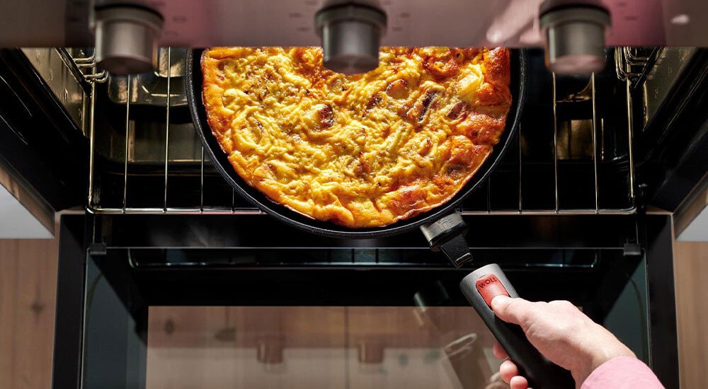 Eine Person holt eine goldbraune Quiche in einer schwarzen Pfanne mit abnehmbarem Griff aus einem offenen Backofen.