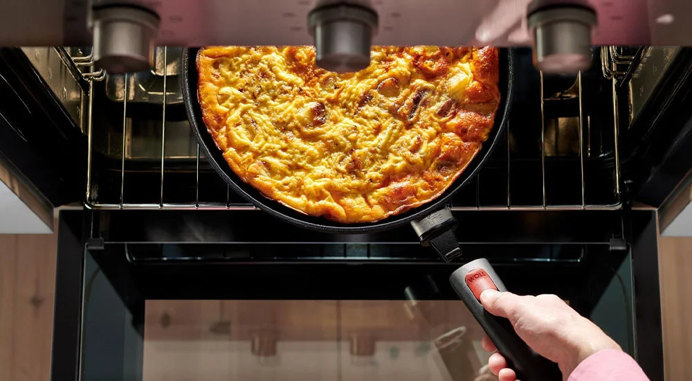 Eine Person holt eine goldbraune Quiche in einer schwarzen Pfanne mit abnehmbarem Griff aus einem offenen Backofen.