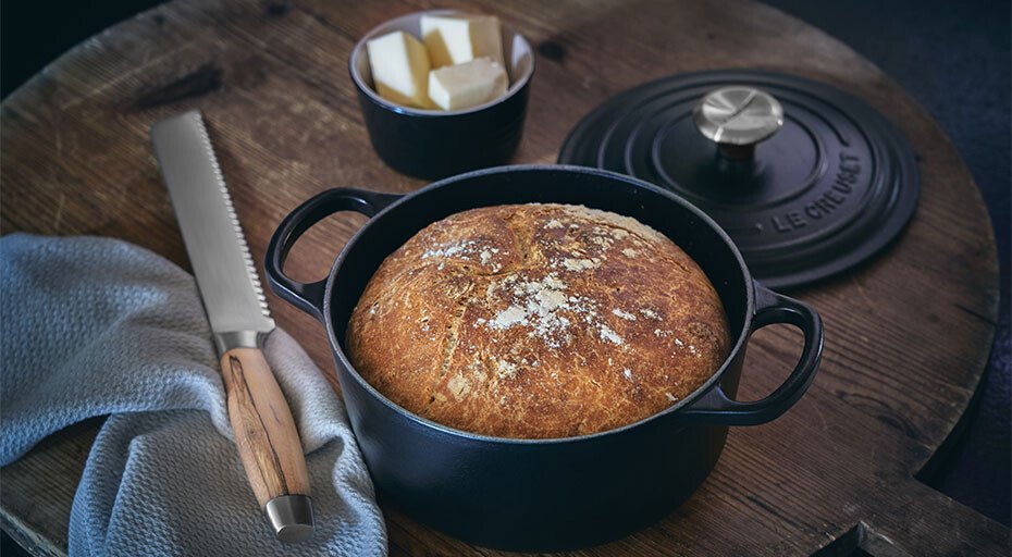 Gusseisen-Bräter von Le Creuset mit frisch gebackenem Brot, daneben Messer und Butterwürfel auf Holztisch.