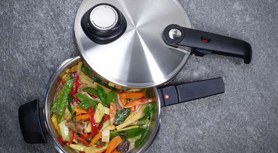 Fissler Vitavit Premium Schnellkochtopf mit geöffnetem Deckel, gefüllt mit buntem Gemüse wie Paprika und Zuckerschoten, auf grauer Oberfläche.