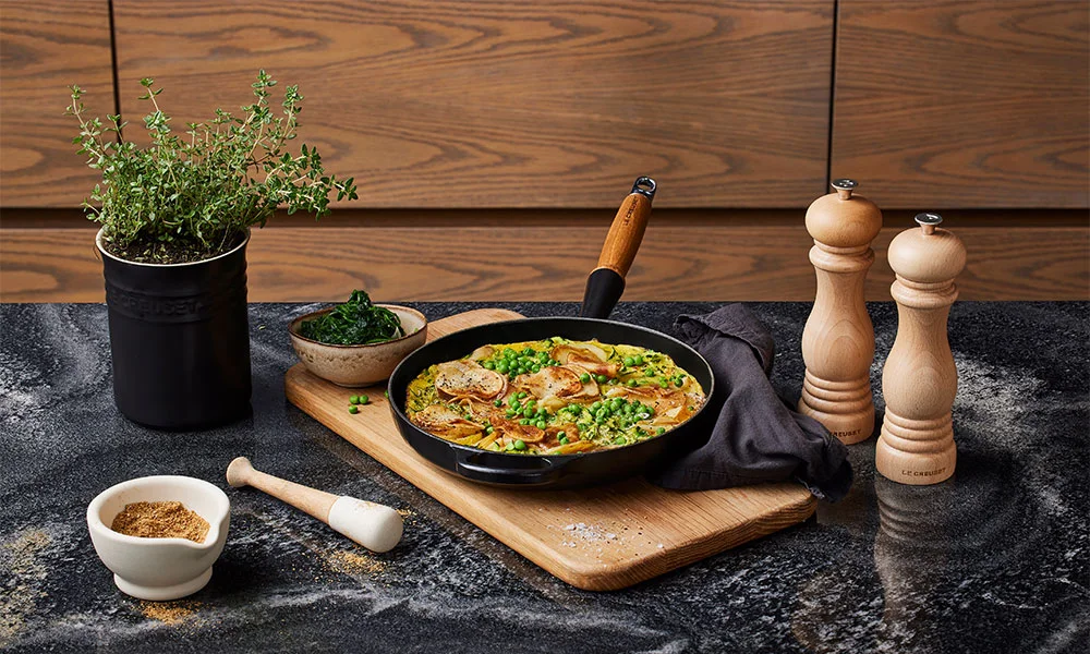Le-Creuset-Pfanne mit Fleisch und Gemüse auf Holzbrett, Pfeffermühlen, Gewürzschale und Kräutertopf auf Marmortisch vor Holzvertäfelung.