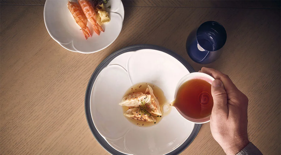 Eine Hand gießt Sauce über Garnelen auf einem Teller der Serie Bloom von Rosenthal, daneben ein Teller mit weiteren Garnelen und ein Glas Wasser.
