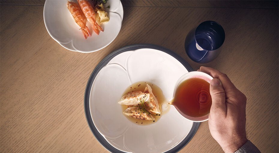 Eine Hand gießt Sauce über Garnelen auf einem Teller der Serie Bloom von Rosenthal, daneben ein Teller mit weiteren Garnelen und ein Glas Wasser.