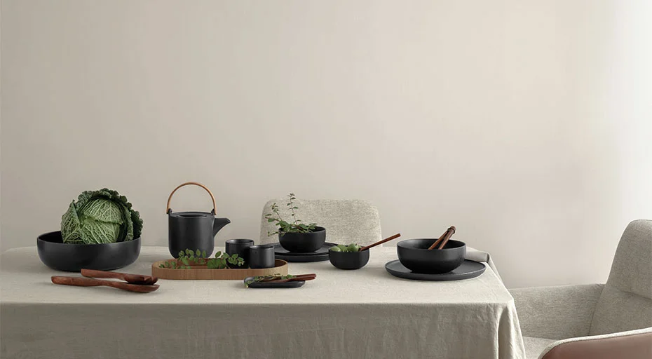 Gedeckter Tisch mit schwarzem Geschirr der Serie Coppa Kuro von ASA, Kräutern und einer Kohlsorte auf hellgrauer Tischdecke vor weißer Wand.