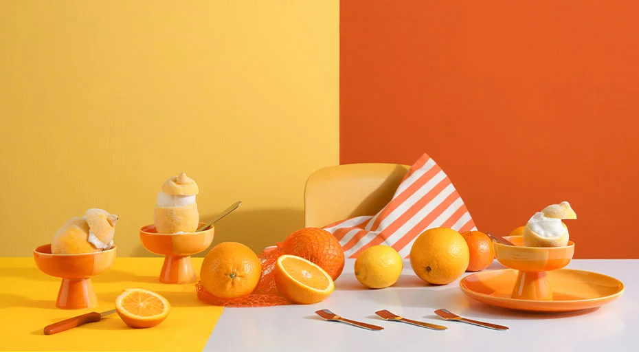 Geschirr der Serie Joy von ASA mit Zitrusfrüchten, bunten Dessertschälchen und orangefarbenen Accessoires vor farbigem Hintergrund.