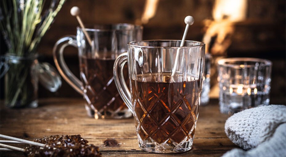 Zwei befüllte Teegläser, verziert mit Kandissticks, stehen auf Holztisch, umgeben von Kerzen und Textilien.