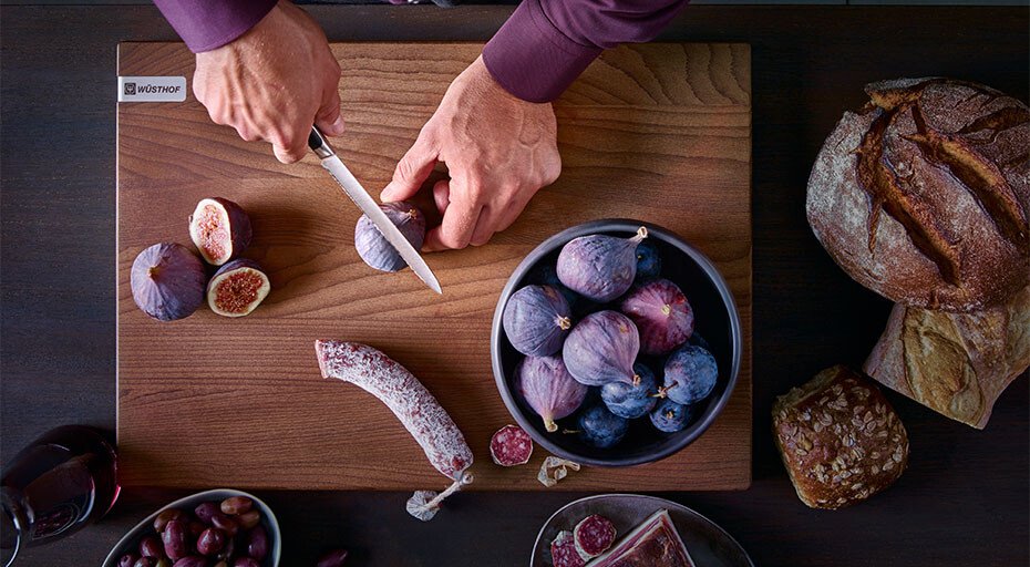 Person schneidet mit einem Messer der Serie Classic Ikon von Wüsthof Feigen auf Holzbrett, umgeben von Brot, Salami, und Schale mit Feigen und Pflaumen.