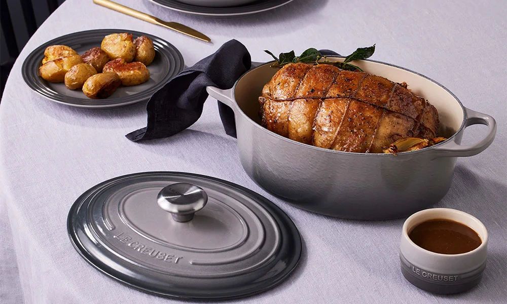 Le Creuset Bräter mit Braten, Teller mit Kartoffeln, Teller mit Bratensaft. Tisch mit weißem Tuch, Deckel und Messer.