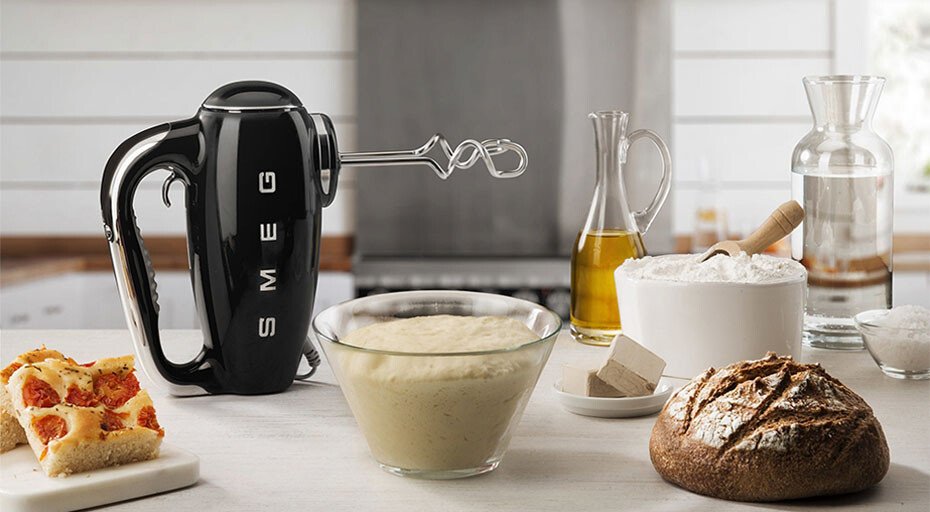 Schwarzer Handmixer von SMEG auf Küchentheke mit Teigschüssel, Mehl, Brot, Hefewürfel und Olivenöl im Hintergrund.