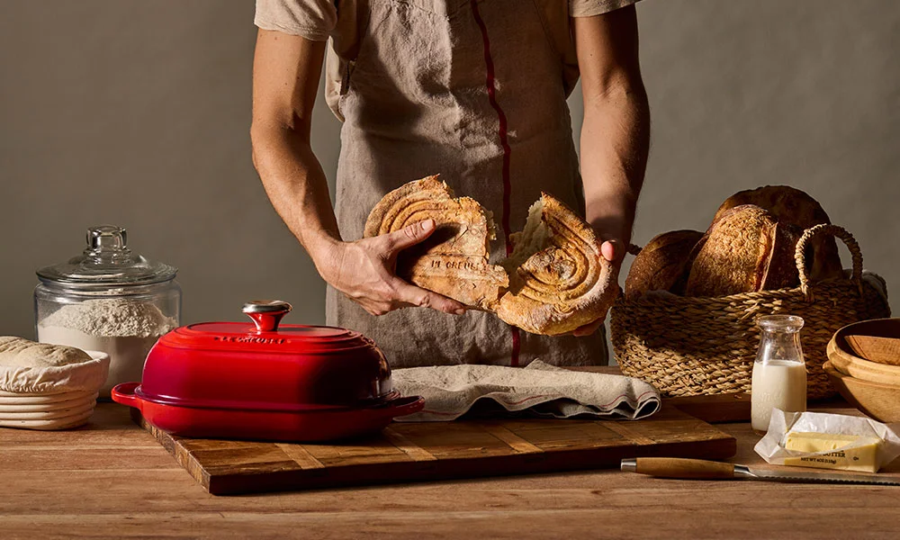 Person schneidet handgefertigtes Brot, umgeben von Backzutaten und Utensilien auf einem Holztisch.