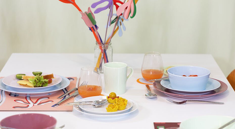 Ein gedeckter Tisch mit buntem Geschirr der Serie Tric Blue Fog von Thomas, Gläsern und Vasen mit dekorativen Zweigen.