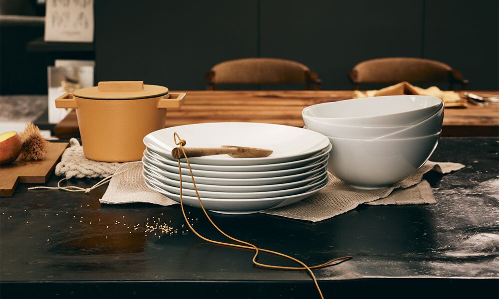 Gestapelte weiße Teller und Schalen auf Tisch, mit Topf und Holzlöffel. Küchenutensilien in moderner Umgebung.