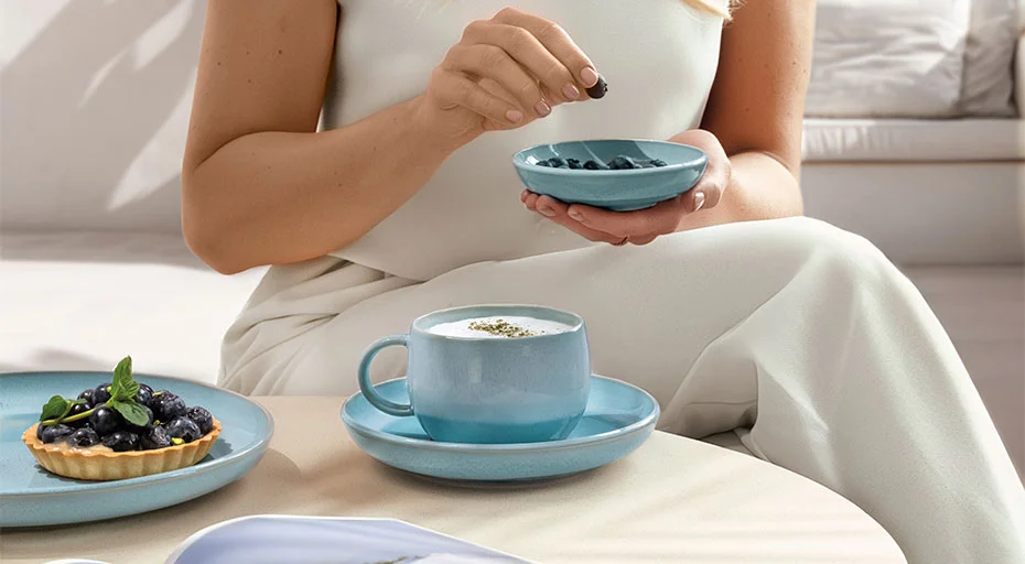 Geschirr von Villeroy &amp; Boch Perlemor Aqua: Person in weißer Kleidung hält Schale mit Blaubeeren, daneben ein Cappuccino in blauer Tasse und Blaubeertörtchen auf Tisch.